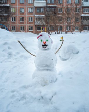 A cheerful snowman stands in the courtyard of the house, snowdrifts on the background
