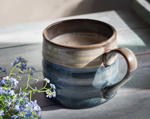 Büyük bir seramik mavi-gri bardakta kapuçino ahşap bir masada, küçük bir buket unutma-beni-nots