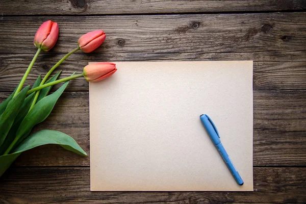 Three red Tulip and a piece of paper with a pen on a brown wooden background — Stock Photo, Image