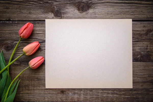 Три красных тюльпана и лист бумаги на коричневом деревянном фоне — стоковое фото