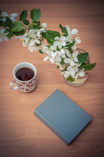 Un livre fermé sur la table avec une branche de fleurs de pomme et une tasse de thé — Photo