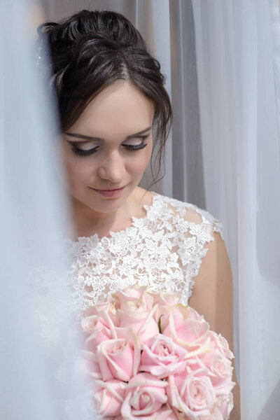 Красивая невеста у окна смотрит на свадебный букет из розовых роз
