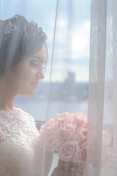 Beautiful bride at the window looking at a wedding bouquet of pink roses