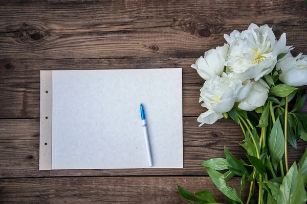 Un foglio di carta con una penna e alcune peonie bianche su sfondo di legno — Foto Stock