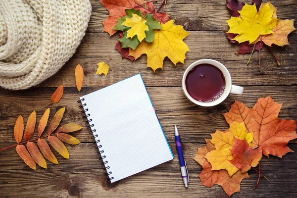 Autumn picture of yellow leaves, a cup of tea, a scarf and a piece of paper with pen on wooden background