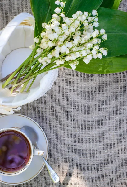 Frühlingsstrauß der Maiglöckchen in einem weißen Korb und einer Tasse Tee — Stockfoto