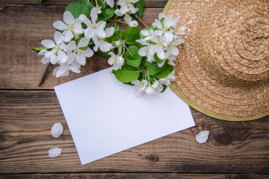 A branch of a blooming apple tree with white flowers, a sheet of paper and a straw hat on a wooden background, with a copy space