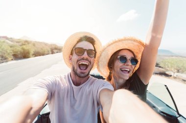Mutlu çift, üstü açık bir arabayla yolculuk ederken selfie çekiyor.