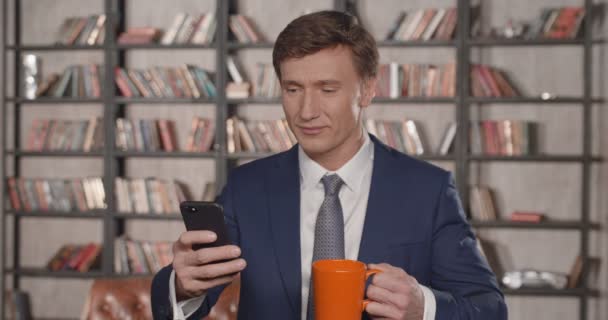 Slow Motion portret van succesvolle aantrekkelijke zakenman typen van berichten in Smartphone en drinken koffie op kantoor glimlachen. Zakenman serie. 4k 4096 x 2160. — Stockvideo