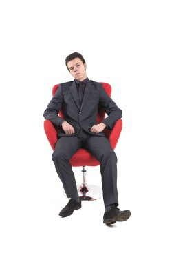 İş takım elbiseli bir genç adam beyaz arka plan üzerinde izole bir kırmızı koltuğa oturur