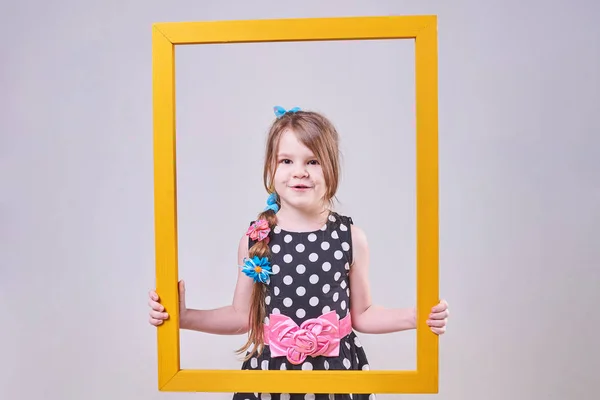 Schönes kleines Mädchen mit einem lustigen Gesichtsausdruck, das einen gelben Rahmen hält — Stockfoto