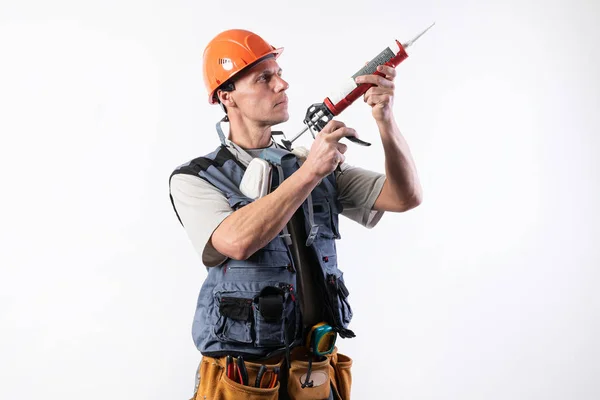 Un constructor en un casco, con silicona en una pistola de montaje. Aguanta en las manos. Sobre un fondo claro . — Foto de Stock