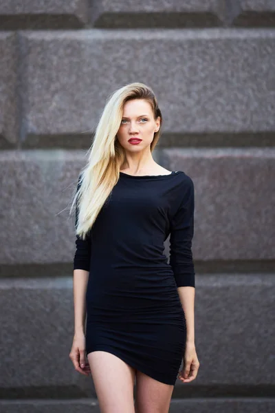 Atractiva rubia con un vestido negro. En pleno crecimiento. Camina con un andar femenino. Sobre el fondo de un muro de piedra . —  Fotos de Stock