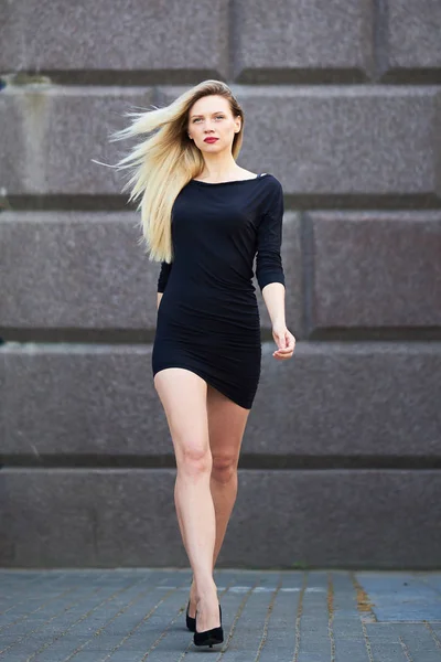 Aantrekkelijk blond in een zwarte jurk. In volle groei. Hij loopt met een vrouwelijke gang. Tegen de achtergrond van een stenen muur. — Stockfoto