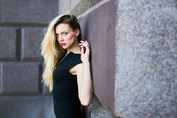 Vacker blond flicka i svart kortklänning. Stående med ryggen mot en stenmur. — Stockfoto