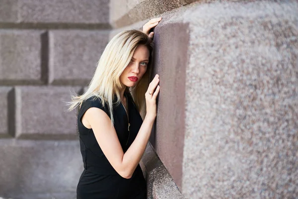 Bella ragazza bionda in un abito corto nero. In piedi con la schiena contro un muro di pietra . — Foto Stock