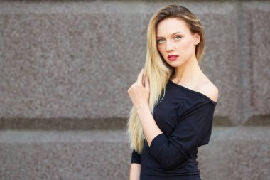 Siyah elbiseli çekici sarışın. Büyürken. Kadınsı bir yürüyüşle yürüyor. Taş bir duvarın arkasında.