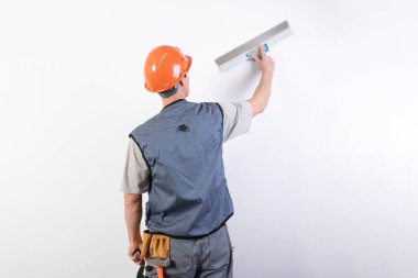 İnşaatçı bir spatulayla duvarı düzleştirir. İş kıyafetleri ve şapkayla. Açık gri bir arkaplanda.