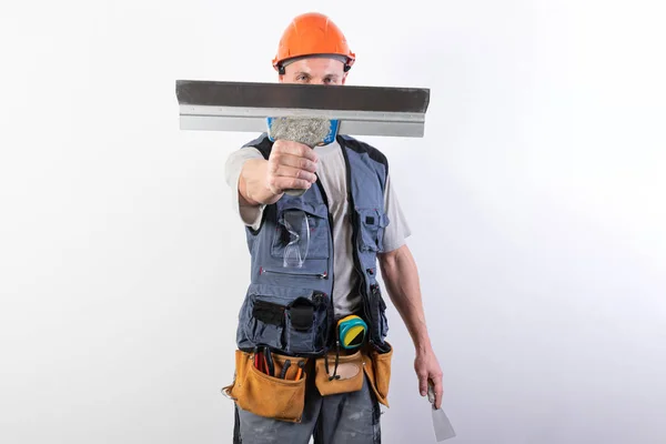 O construtor esconde o rosto atrás de uma espátula. Em roupas de trabalho e chapéu duro. Em um fundo cinza claro . — Fotografia de Stock