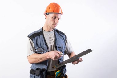 İnşaatçı bir spatulayı temizliyor. İş kıyafetleri ve şapkayla. Açık gri bir arkaplanda.