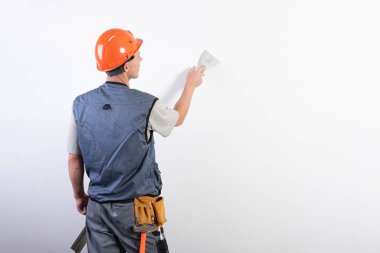 İnşaatçı bir spatulayla duvarı düzleştirir. İş kıyafetleri ve şapkayla. Açık gri bir arkaplanda.