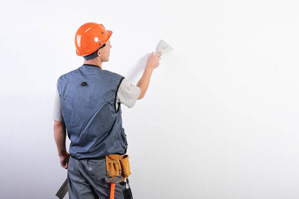 The builder smoothes the wall with a spatula. In work clothes and hard hat. On a light gray background.