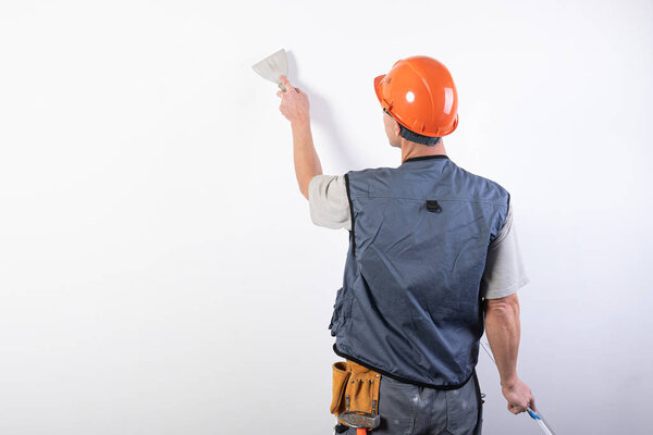 The builder smoothes the wall with a spatula. In work clothes and hard hat. On a light gray background.