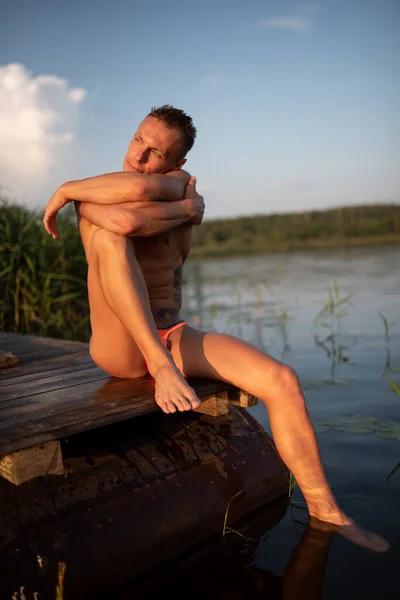 Uomo atletico sul lago. Si siede sul molo . — Foto Stock
