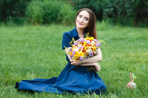 Mulher em um vestido azul está sentado em um prado com flores e beber suco — Fotografia de Stock