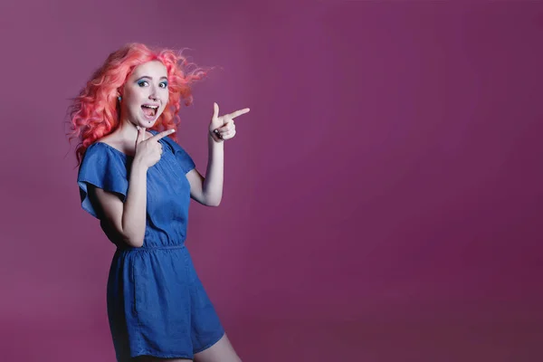 Όμορφη κοπέλα με ροζ μαλλιά σε μπλε φόρεμα δείχνει σημάδι σε λιλά φόντο, τοποθετήστε για κείμενο — Φωτογραφία Αρχείου