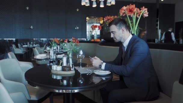 Empresario se sienta en una mesa en un restaurante y utiliza su teléfono inteligente — Vídeos de Stock