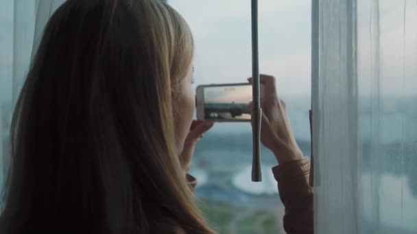 The girl stands in front of the window and shoots the cityscape on a smartphone — Stock Video