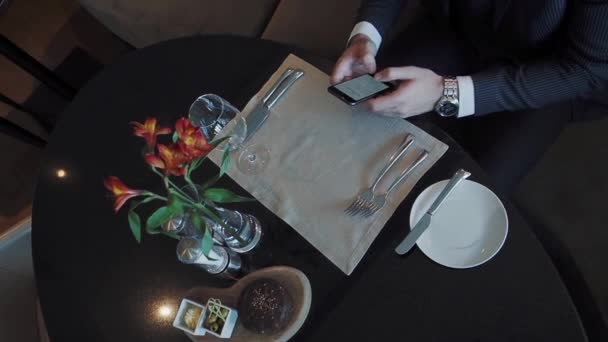 Empresario se sienta en una mesa en un restaurante y utiliza su teléfono inteligente — Vídeos de Stock