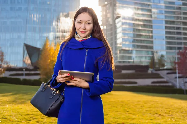 Asian woman holding tablet in hand, standing instanding outdoors behind skyscrapers