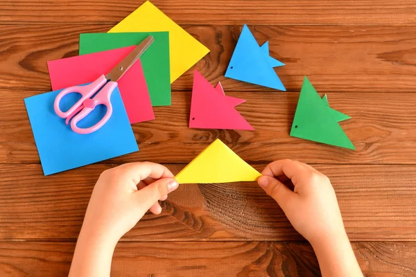 Origami pesci colorati, fogli di carta, forbici. Bambino tiene foglio di carta in mano e fare origami pesce. Tavolo in legno marrone — Foto Stock