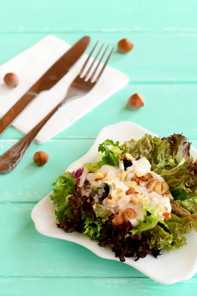 Mix of salad leaves with yogurt and hazelnuts on a plate. Salad is prepared from radicchio, lettuce, iceberg, frieze. Fork, knife, white napkin, raw hazelnuts on a wooden background. Closeup