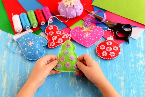 Small child holds felt Christmas tree in his hands. Child shows Christmas crafts. Felt fur-tree, mitten, heart, ball decor on wood background. Flat felt sheets, scissors, sewing set, pins, pincushion — Stock Photo, Image