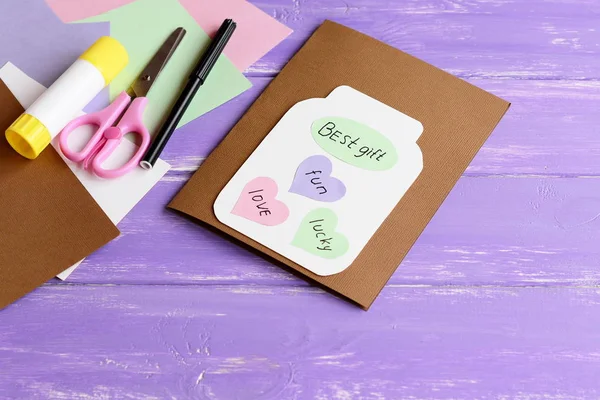 Paper greeting card with jar, colored hearts and wishes. Scissors, colored paper sheets, white cardboard, glue stick, black marker on wooden background. Fun kids art concept — Stock Photo, Image