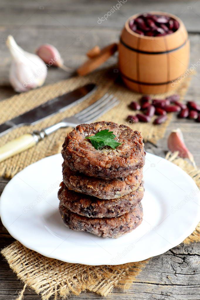 Fried bean cutlets with garlic and spices on a plate. Small decorative barrel with uncooked red beans, garlic, parsley, fork, knife on wooden background. Healthy diet cutlets. Rustic style. Closeup 