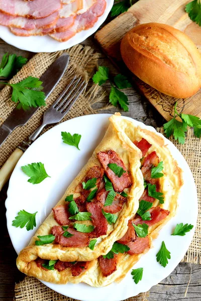 Bacon omeleta recept. Domácí omeleta se slaninou a petrželkou na talíři, plátky slaniny na talíř, rohlík, vidličku, nůž, prkénko, čerstvá petržel větvičky na staré dřevěné pozadí. Pohled shora — Stock fotografie