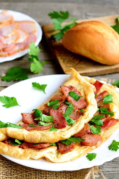 Omelete de bacon caseiro. Deliciosa omelete recheada com bacon e salsa em um prato, fatias de bacon em um prato, pão, garfo, faca, tábua de corte, raminhos de salsa frescos na velha mesa de madeira. Fechar. — Fotografia de Stock