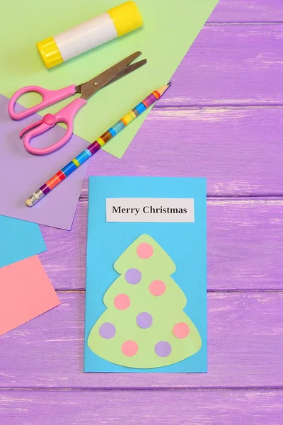 Paper card Merry Christmas. Cute greeting card with paper Christmas tree, scissors, glue stick, pencil, colored paper set on lilac wooden background. Simple children Christmas paper crafts