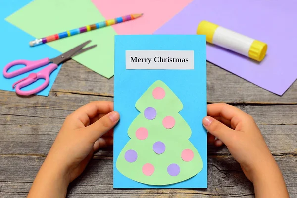 Child holding Christmas card in his hands. Paper greeting card with text Merry Christmas and Christmas tree with ornaments. Simply children winter crafts diy at home or in kindergarten. Greeting cards making. Craft supplies 