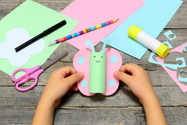 Enfant tenant papillon de papier coloré dans les mains. Enfant montre un bricolage papier amusant. Papeterie sur un vieux fond en bois. Art du papier d'été préscolaire et activités artisanales de colle. Développement des capacités créatives des enfants — Photo