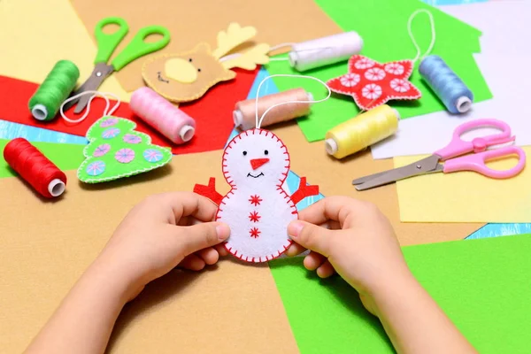 Small child holds a felt Christmas snowman ornament in hands. Child shows Christmas ornament crafts. Beautiful felt Christmas tree ornaments, scissors, thread, needles, felt sheets on table. DIY xmas gifts for kids to make creative. Kids festive diy — Stock Photo, Image