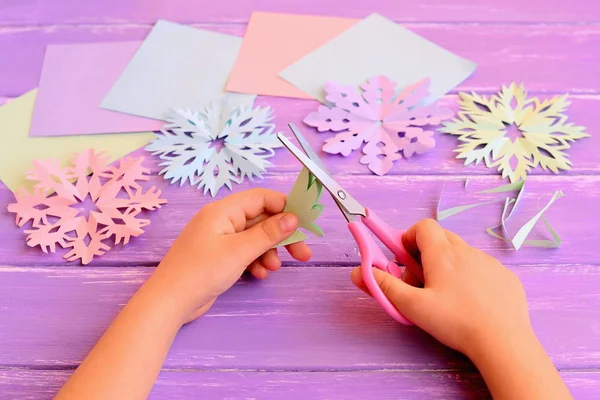 Il bambino taglia un fiocco di neve di carta. Fogli di carta colorati, rosa, verde, viola, blu fiocchi di neve su un tavolo di legno sfondo invernale. Ritagli di carta di Natale idee artigianali. Pratica di taglio delle forbici per bambini a casa o all'asilo — Foto Stock