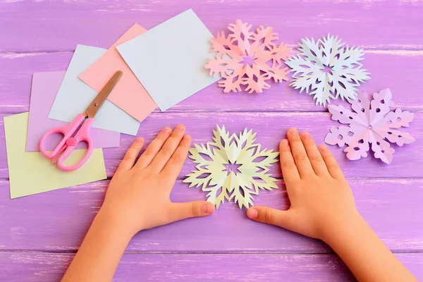 Μικρό παιδί δείχνει χριστουγεννιάτικο χαρτί νιφάδες χιονιού αποκόμματα. Παιδιά χέρια σε λιλά ξύλινο τραπέζι. Όμορφα χρωματιστά νιφάδες χιονιού κομμένα από χαρτί, προμήθειες χειροτεχνίας, ψαλίδι. Εύκολη ιδέα για χειμωνιάτικα παιδιά. Χειμερινή δραστηριότητα για παιδιά στο σπίτι — Φωτογραφία Αρχείου