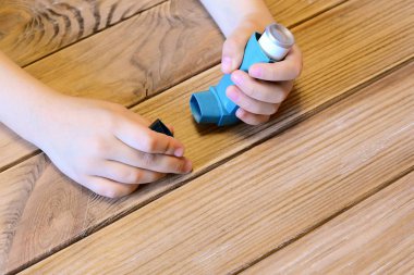 Small kid opens asthma inhaler. Inhalation treatment of respiratory diseases. Allergy and bronchial asthma medication. Old wooden background  clipart