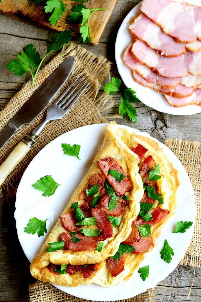 Fried bacon omelette. Omelette stuffed with bacon and parsley on a plate, bacon slices on a plate, fork, knife, cutting board, parsley sprigs on wooden background. Homemade eggs breakfast. Top view 