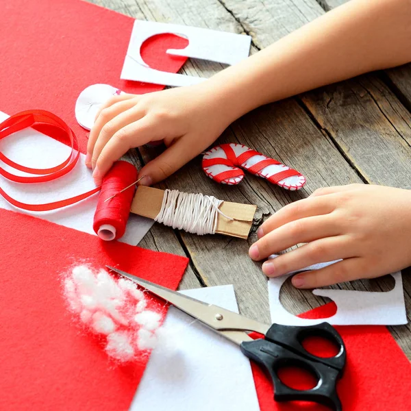 Çocuklar ellerinde eski ahşap masa. El yapımı keçe Noel candy süsleme, keçe çarşafları ve hurda, makas, kırmızı iplik, iğne, kağıt şablonu, pin. Bebek işyeri. Çocuklar Noel sanat projesi — Stok fotoğraf